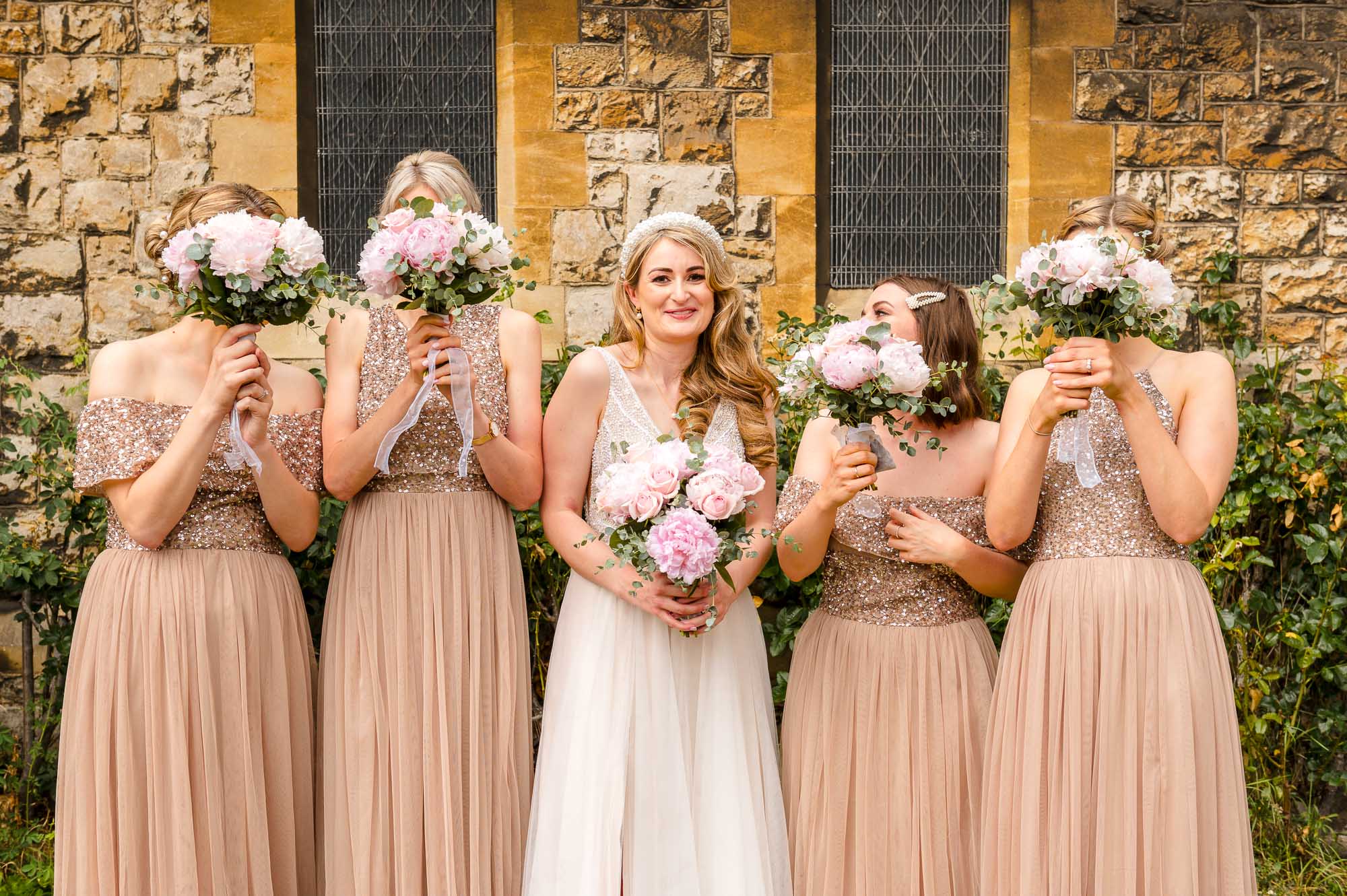 Fun photograph for inspiration of the bride and bridesmaids covering their faces with bouquets in the grounds of St John the Baptist Church in Eltham, London