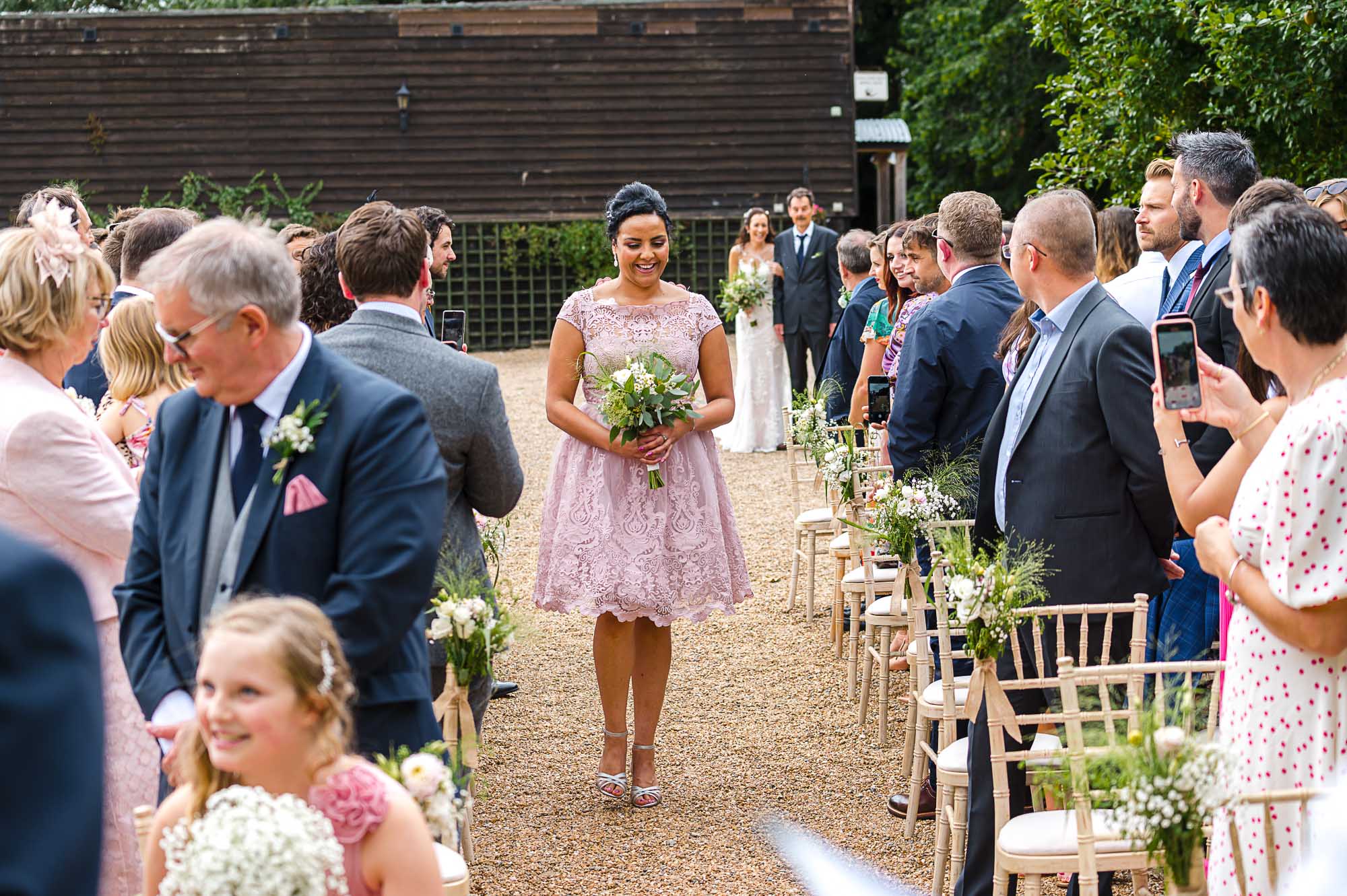 Maid of honor walking down the aisle with bride and her father in the background for the outdoor wedding ceremony at The Pavilion at Swallows Oast wedding venue in Ticehurst, East Sussex