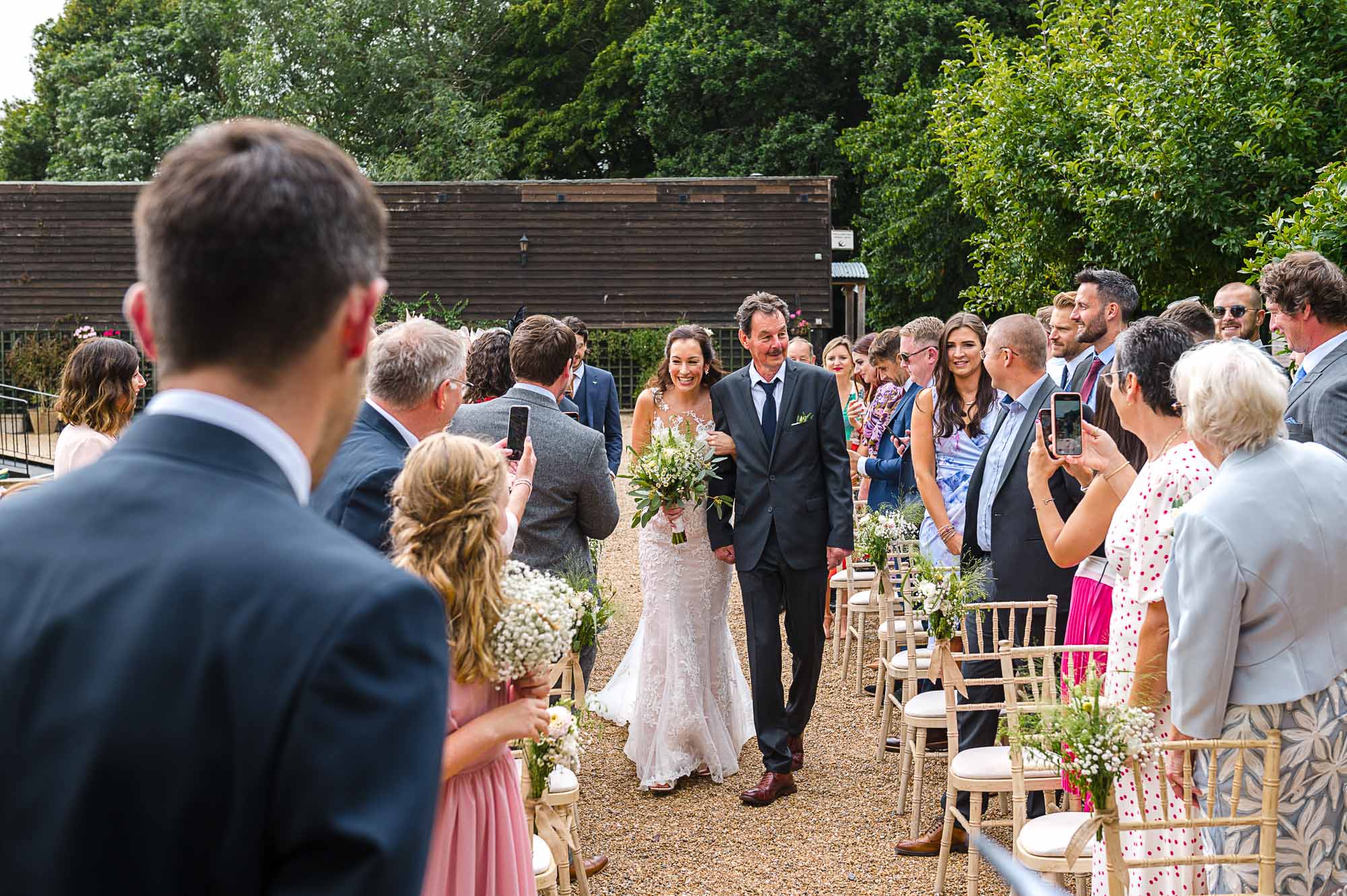 Bride walking down the aisle with her father heading towards The Pavilion for the outdoor ceremony at Swallows Oast wedding venue in Ticehurst, East Sussex