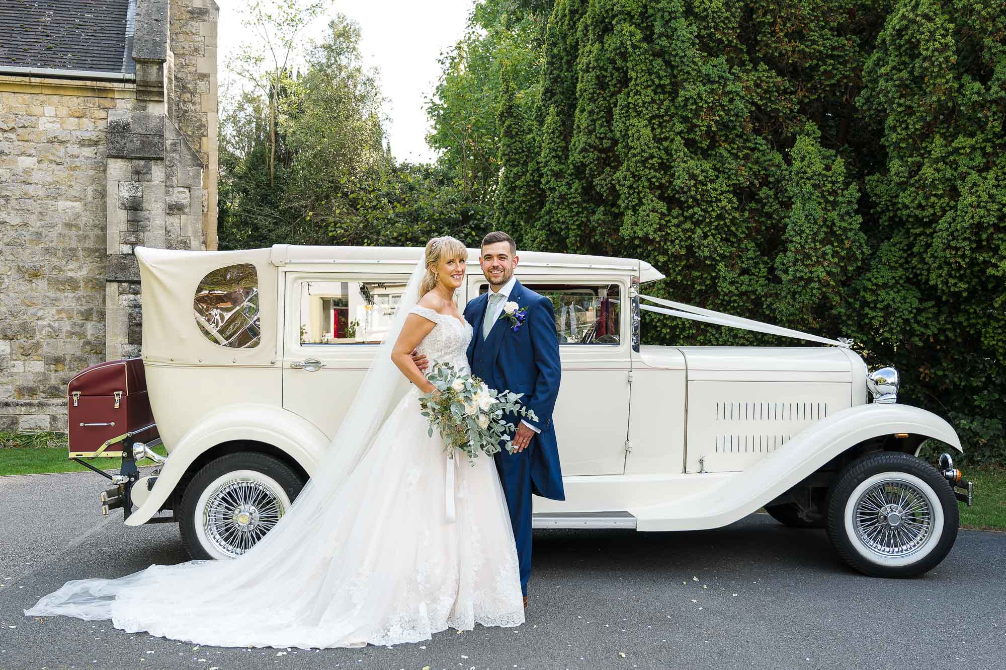 Bride and groom portrait in front of their vintage wedding car in the grounds of St John the Evangelist Church, Bexley, Kent